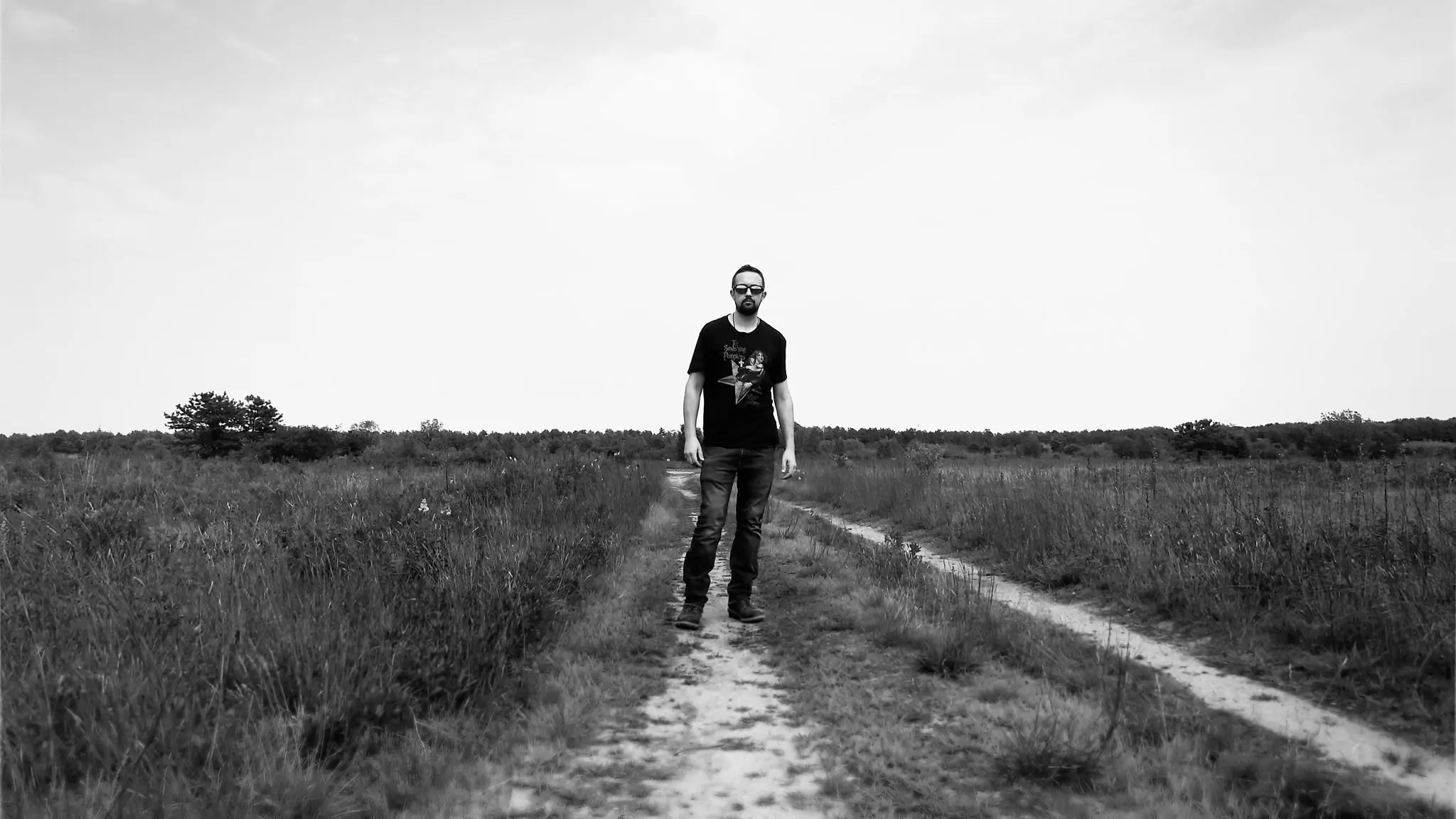 Black-and-white environmental portrait of Thomas Stirling standing on a sandy path amid wide-open coastal barrens beneath a vast sky